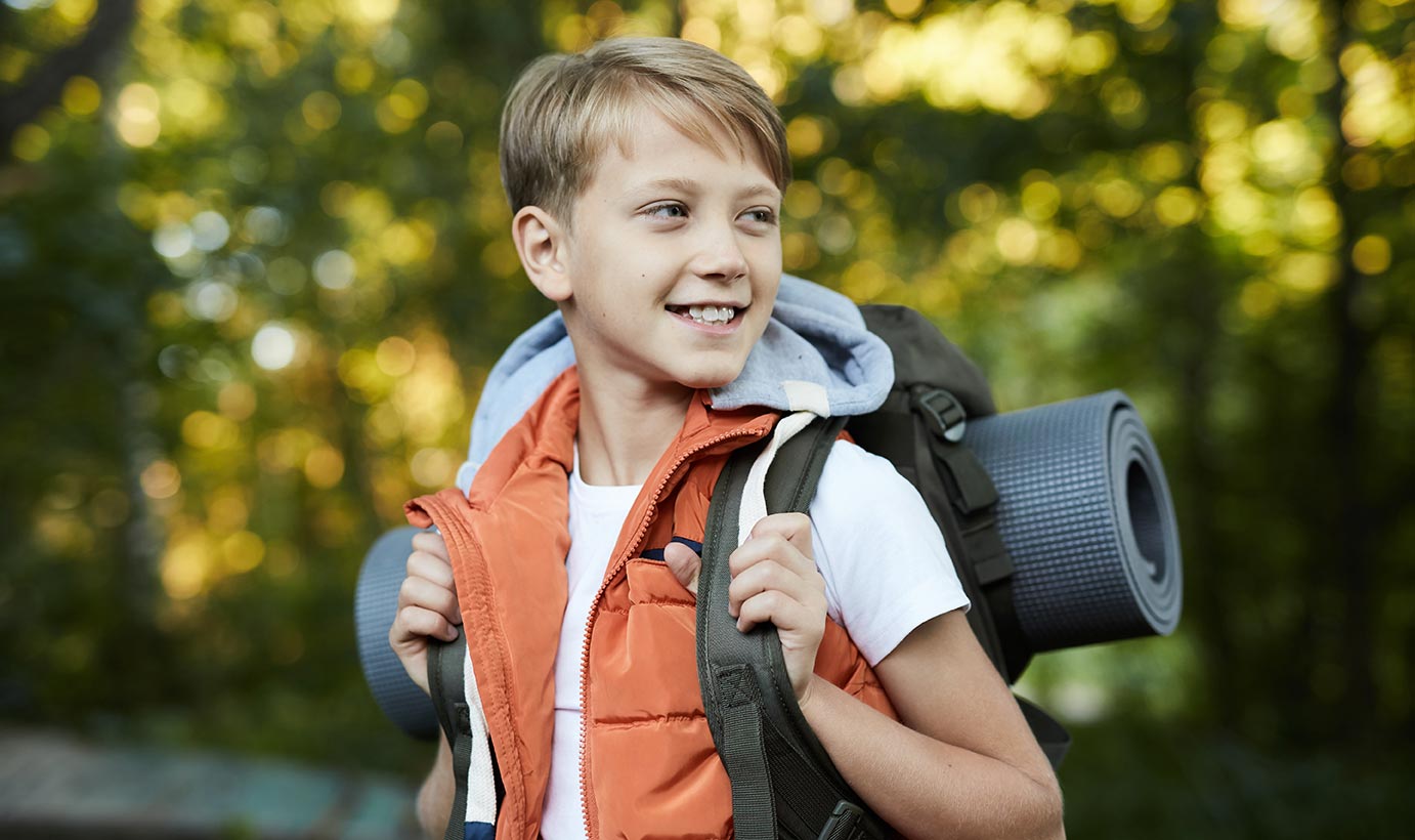 A young boy with healthy teeth wears an orange vest and smiles happily during a camping trip.